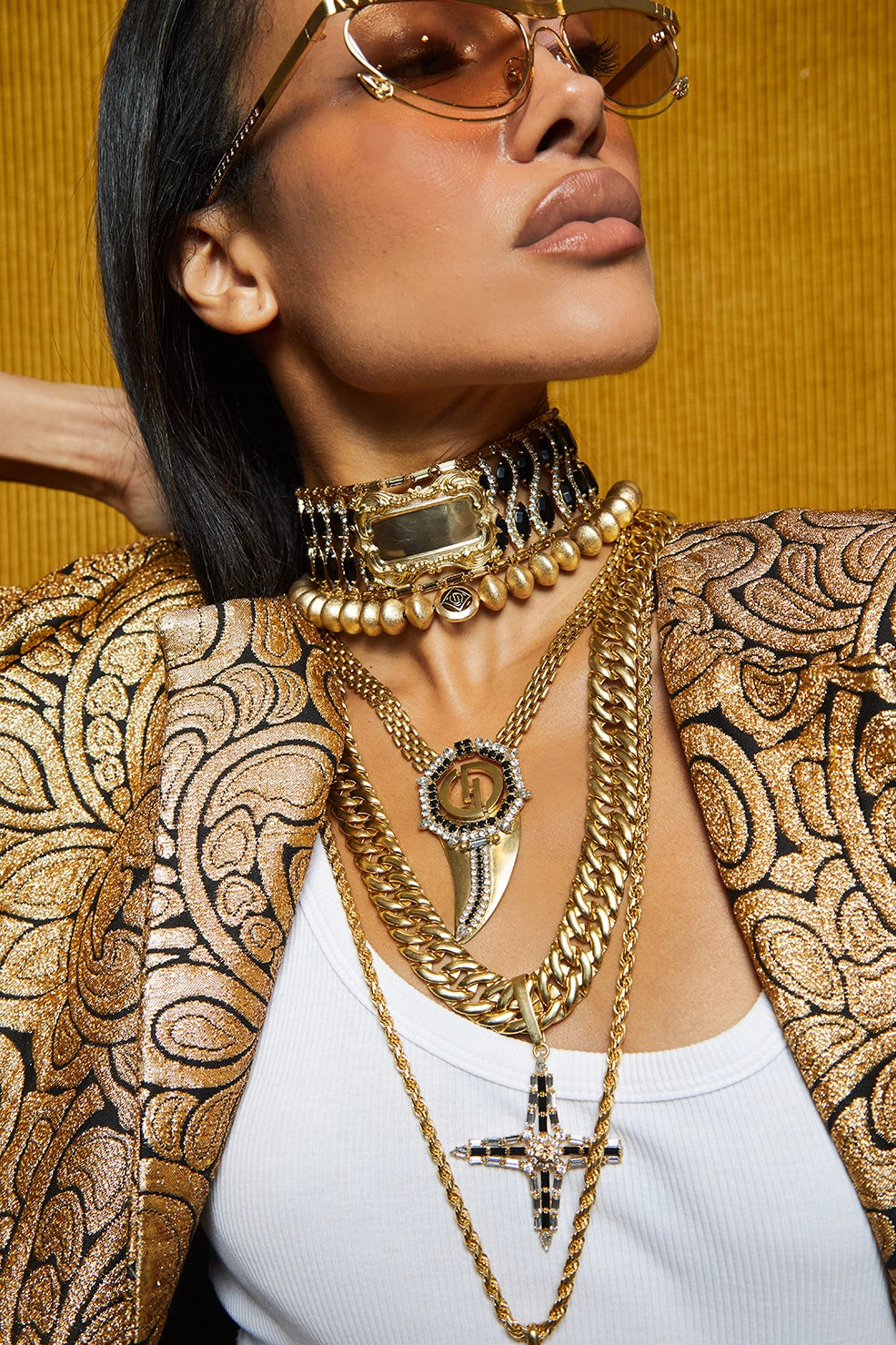 A close-up lifestyle shot of a model wearing an assortment of layered gold necklaces, including a cross pendant, a fang-shaped pendant, and a beaded choker, styled with a gold patterned blazer and sunglasses against a textured gold background.