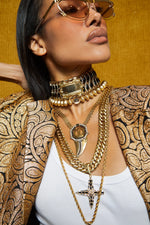 A close-up lifestyle shot of a model wearing an assortment of layered gold necklaces, including a cross pendant, a fang-shaped pendant, and a beaded choker, styled with a gold patterned blazer and sunglasses against a textured gold background.