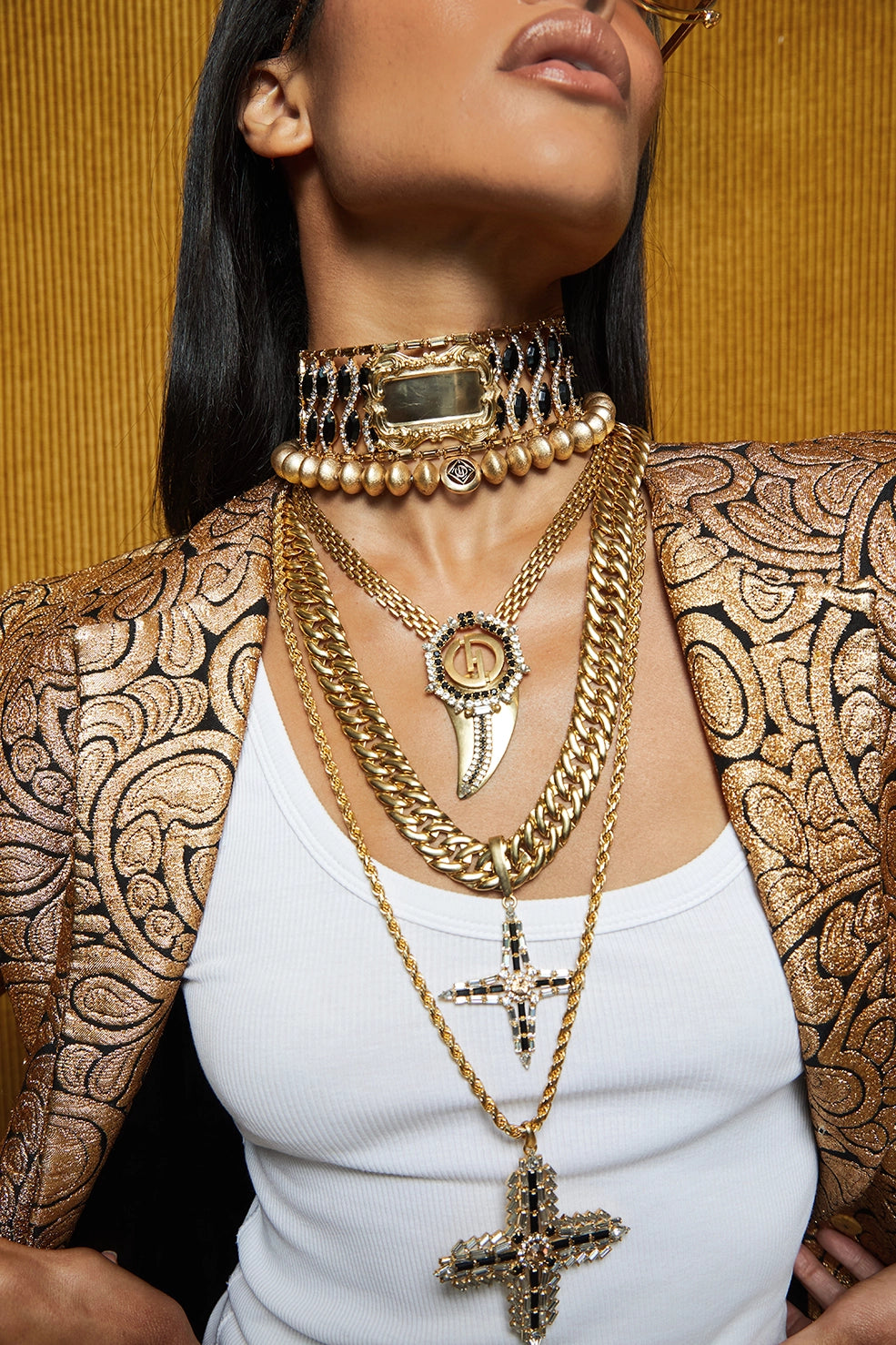 A close-up of a model wearing multiple layered gold necklaces, including a choker, chain necklaces, and a cross pendant, styled with a patterned gold and brown blazer over a white top against a textured gold background.