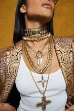 A close-up of a model wearing multiple layered gold necklaces, including a choker, chain necklaces, and a cross pendant, styled with a patterned gold and brown blazer over a white top against a textured gold background.