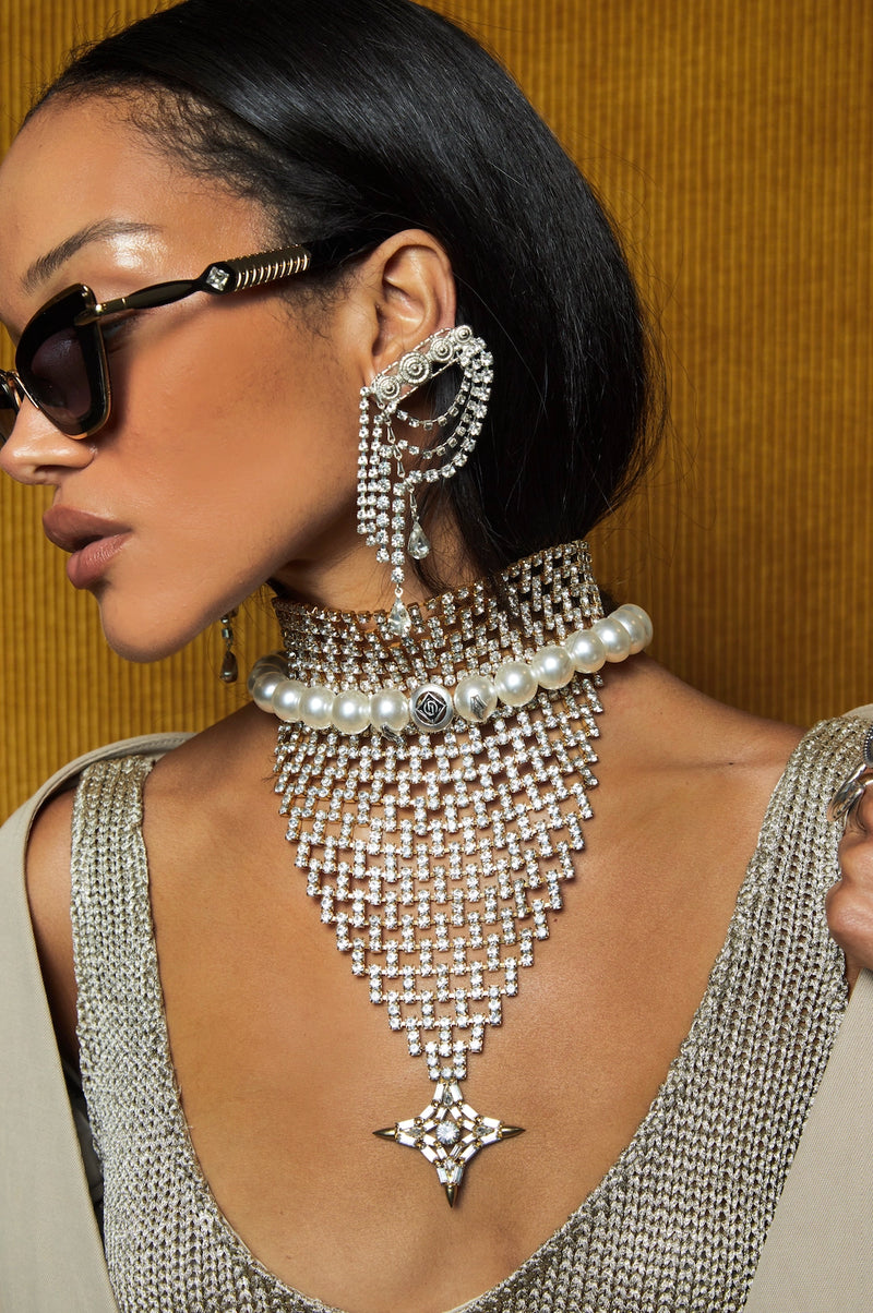 A close-up lifestyle shot of a model wearing an elaborate DYLANLEX silver-toned rhinestone choker necklace with a long cascading fringe and a matching large drop earring, paired with a silver chain-strap top against a textured mustard yellow background.